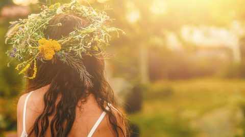 Ansicht einer Frau mit Blütenkranz auf dem Kopf, in Sommerkleid, steht auf einer Wiese